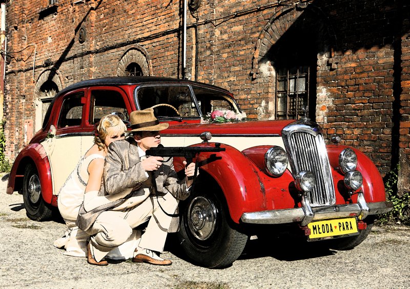 Fotografia ślubna - sesja plenerowa stylizowana na lata 40 gangsterskie - para młoda za samochodem z karabinem